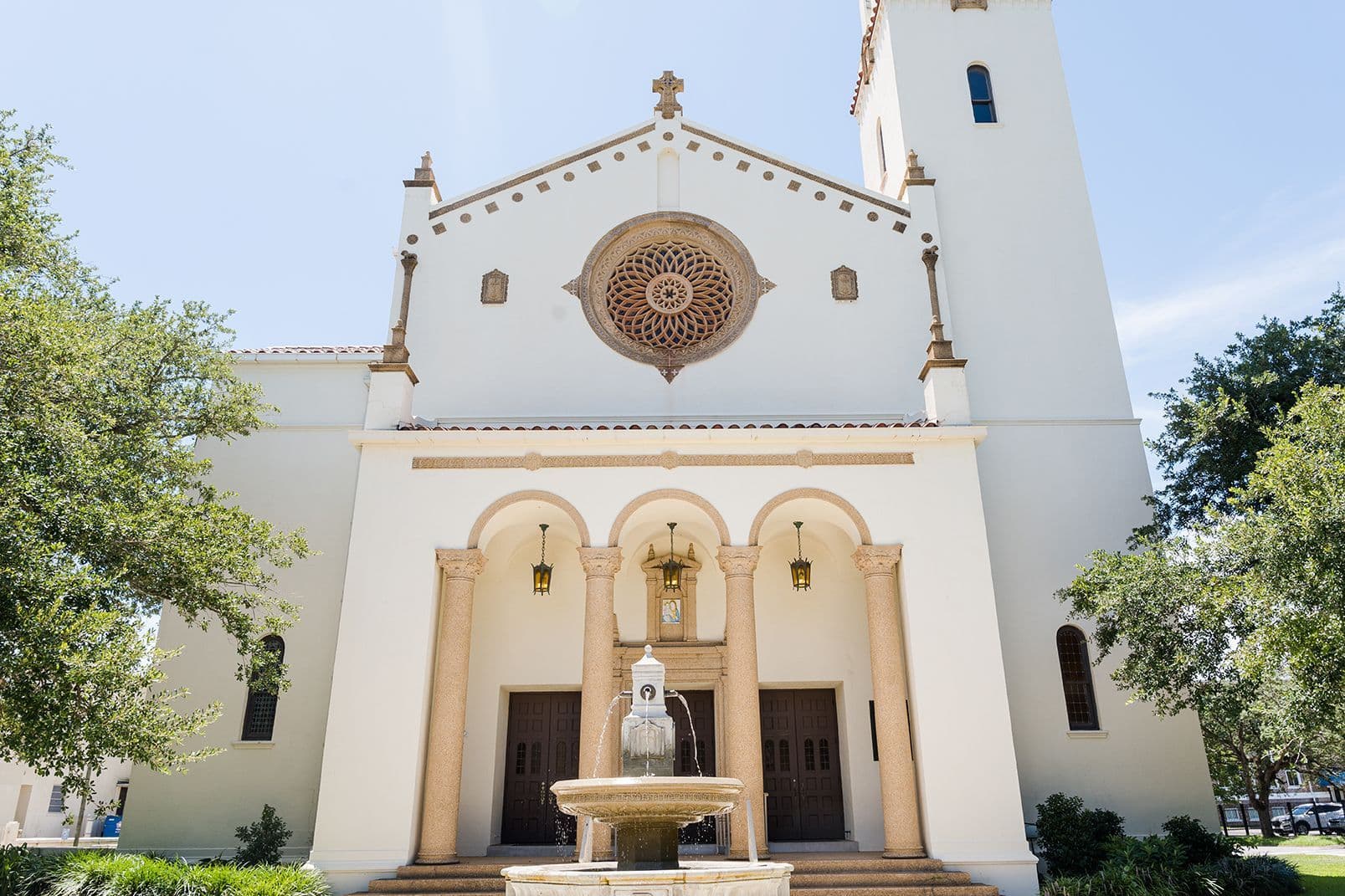 The facade of St. Mary's Catholic Church in Mobile, Alabama — its whitewashed Spanish Colonial front, rose window, and arched entry portico, with the parish fountain in the foreground.