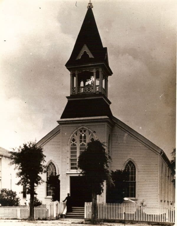 Archival photograph of St. Mary's first church in Mobile, Alabama — a white frame building with a steep steeple and open bell tower, dedicated in 1868 and pictured behind a picket fence.